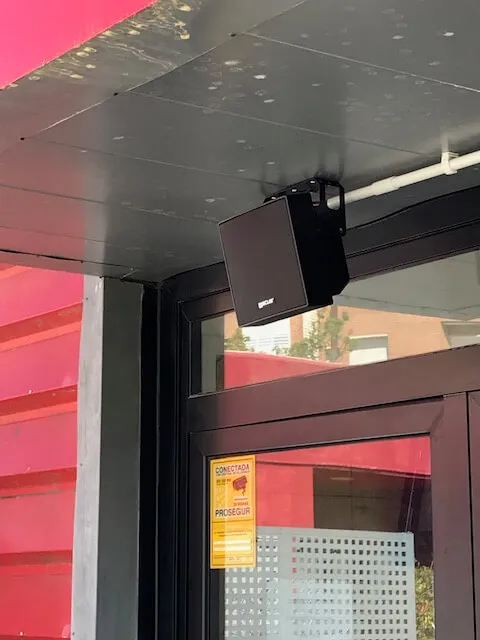 Un altavoz negro está montado en el techo, encima de una puerta de cristal, junto a una señal de seguridad amarilla en la puerta.
