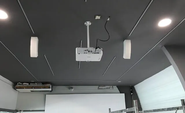 Proyector montado en el techo con cableado a la vista y dos altavoces instalados a cada lado, encima de una pantalla de proyección en una sala moderna.
