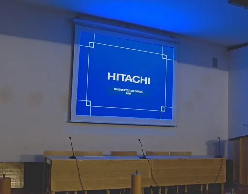 Una sala de conferencias con una pantalla de proyector que muestra la palabra "HITACHI" y varias sillas vacías y micrófonos dispuestos en una larga mesa.