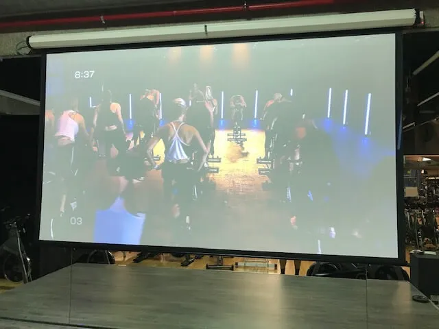 Una pantalla proyectada muestra una clase grupal de ciclismo indoor con los participantes en bicicletas estáticas frente a un instructor en una sala poco iluminada.