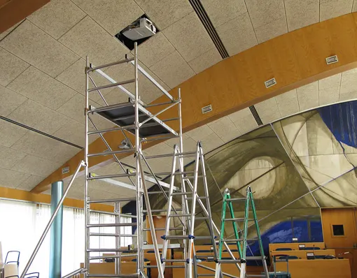 Un andamio metálico y una escalera se colocan bajo un proyector instalado en el techo de una gran sala con modernas obras de arte en la pared e hileras de sillas.