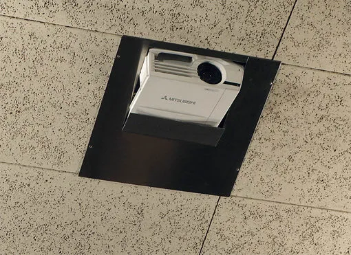Un proyector Mitsubishi montado en el techo está instalado en un marco negro entre placas acústicas blancas.