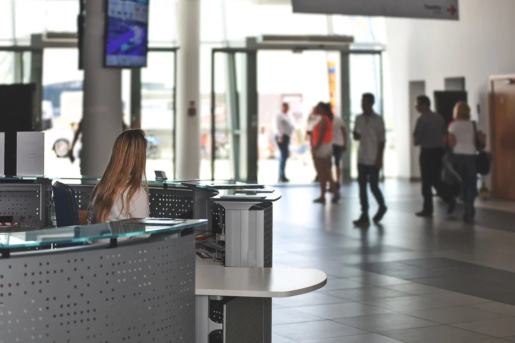 Una recepcionista se sienta en el mostrador de un luminoso y moderno vestíbulo de un edificio equipado con instalaciones multimedia, mientras varias personas entran y salen por las puertas de cristal del fondo.