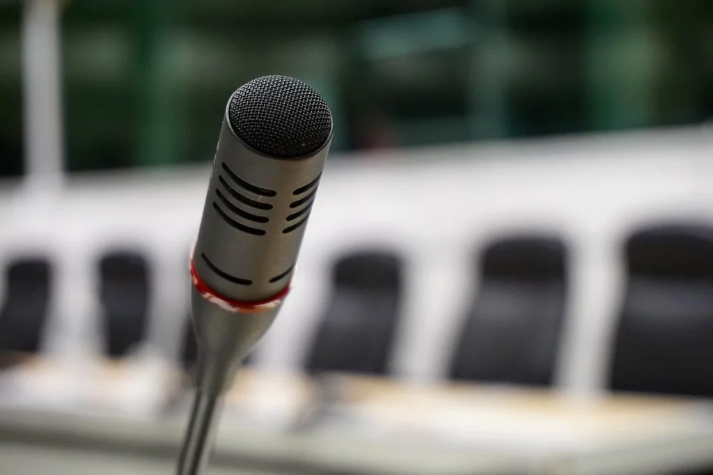 Primer plano de un micrófono con sillas vacías y una mesa de conferencias difuminada al fondo, mostrando modernas instalaciones audiovisuales.