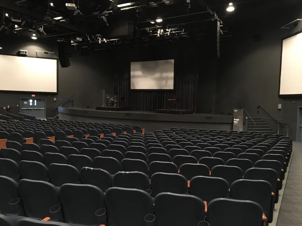 Un auditorio vacío con filas de asientos oscuros frente a un escenario, flanqueado por dos grandes pantallas de proyección, iluminadas por focos cenitales -ideal para instalaciones audiovisuales de última generación-.