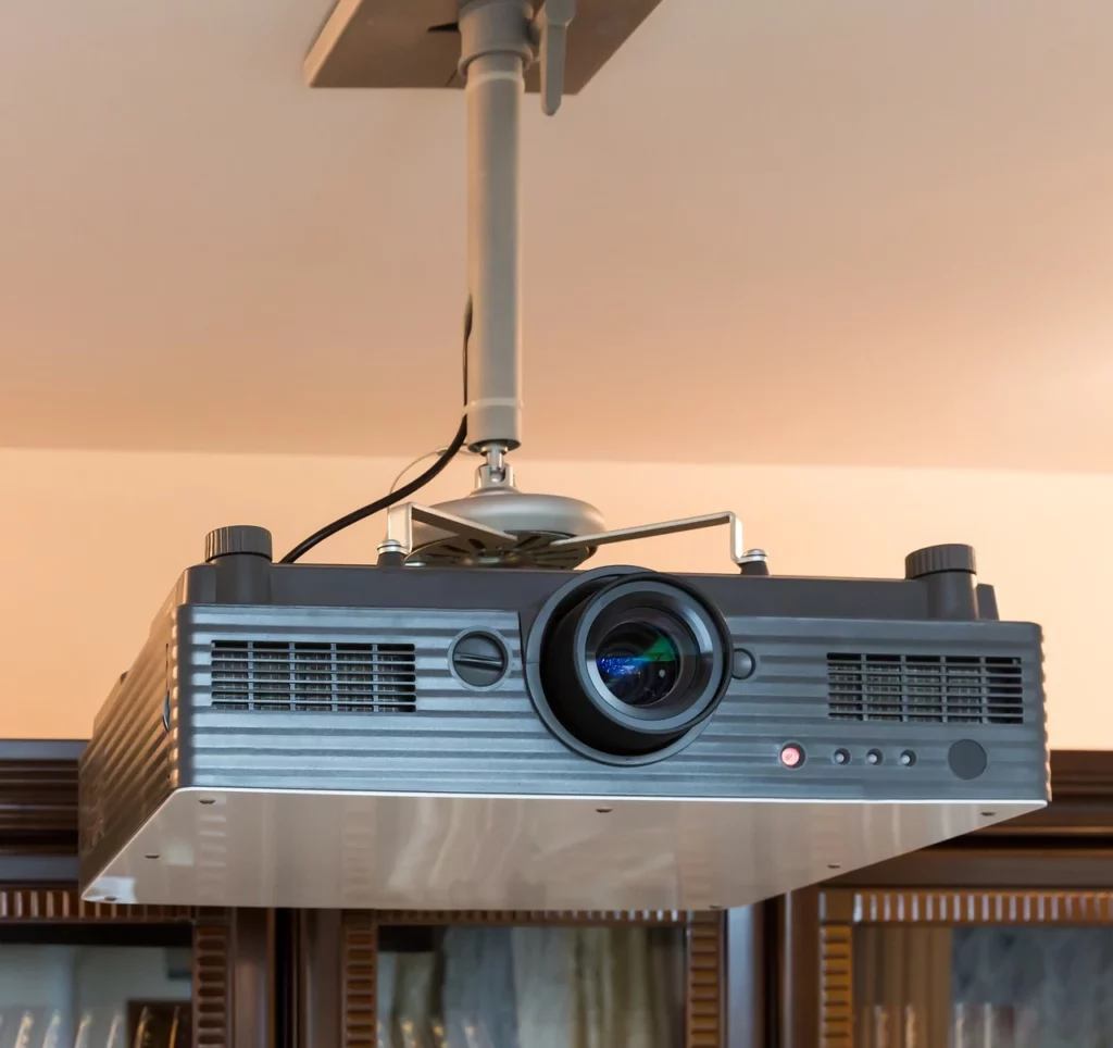 Proyector montado en el techo con una gran lente y mandos de control, colocado en un entorno interior con estanterías y armarios, perfecto para instalaciones audiovisuales que requieren una instalación profesional de proyectores.